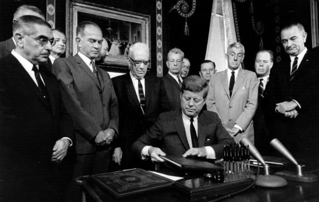 President Kennedy signs the nuclear test ban treaty for the United States watched by a committee of senators, Vice-President Lyndon Baines Johnson and Foreign secretary, Dean Rusk. 
