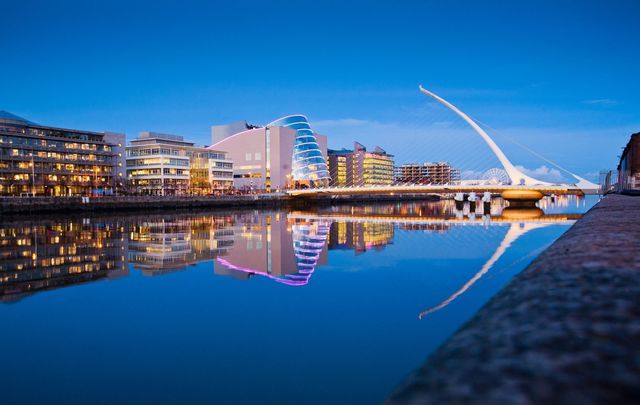 Samuel Beckett Bridge and Convention Center and surrounding buildings on a blue evening with buildings reflected in River Liffey.\n