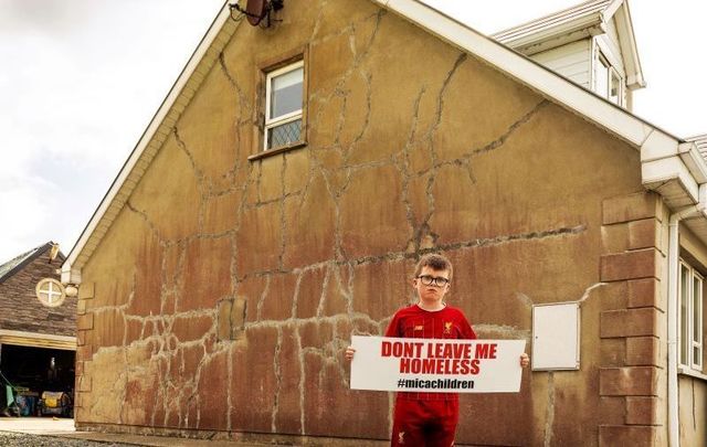 A child stands outside of his home that has cracks in its walls.