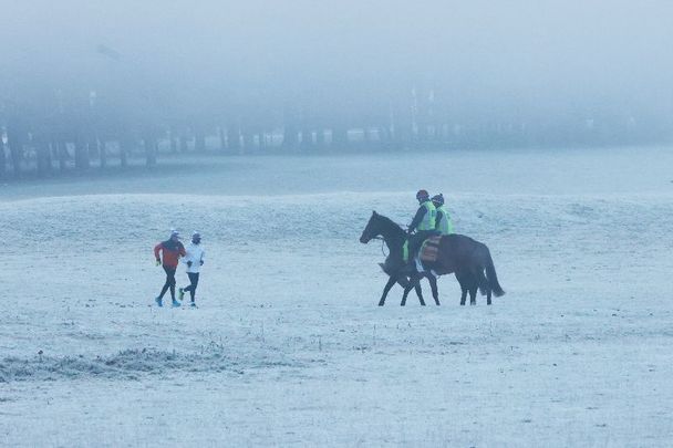 December 9, 2022: Race horses and riders shrouded in fog, with frost and and snow underfoot, encounter two runners, as they return from a morning training run on the Curragh Plains in County Kildare.