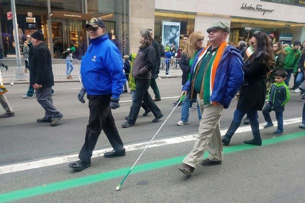 Daniel Ryan Howarth, who is visually impaired, follows the green line up Fifth Avenue in the NYC St. Patrick’s Day Parade. Note the white mobility cane he is using sports green accents in honor of the March 17th celebrations.