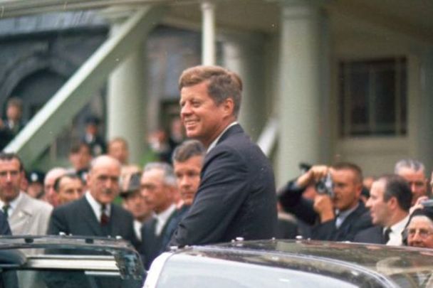 June 28, 1963: President John F. Kennedy stands in the open Presidential limousine as it sits parked in Dublin, Ireland; a crowd looks on. 