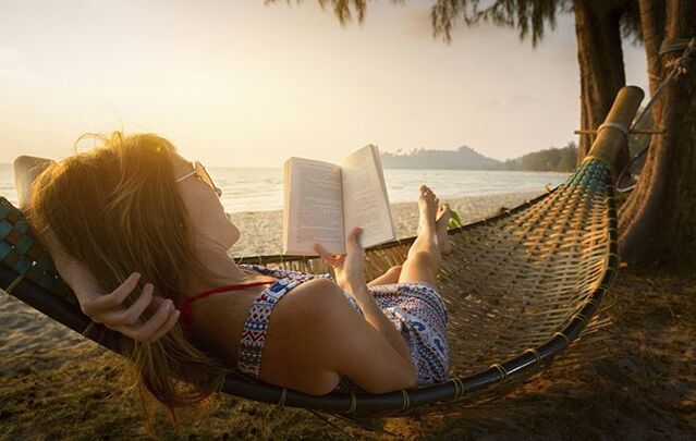 A woman in a hammock reads a book by the beach.