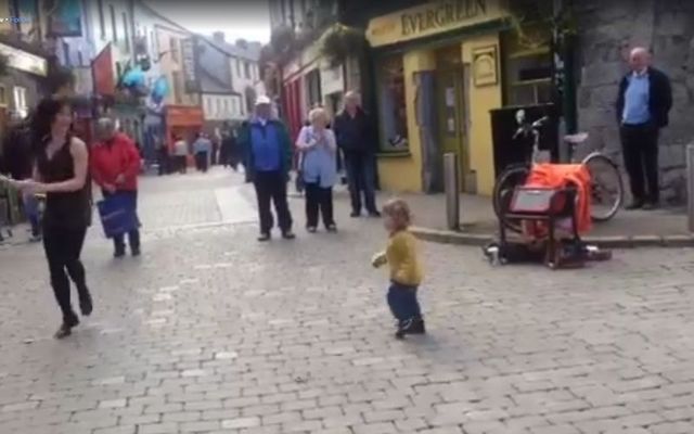 Two-year-old girl tries to copy the steps of an Irish dancer in Galway.  