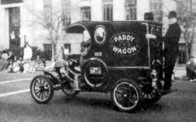 Black-and-white photo of a policeman standing on the back of vehicle, which has the words \"Paddy Wagon.\"