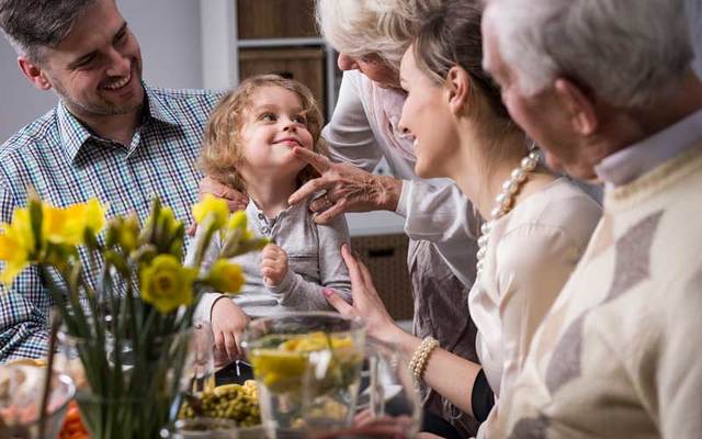 A happy three-generation family smile at a young child around a dinner table.