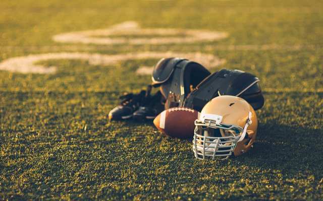 A Gold American Football helmet sits with a football on a football playing field. 