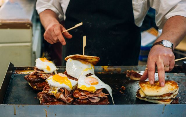 Close view of the cook preparing some sandwiches with bacon, sausages, and fried eggs, on the griddle.