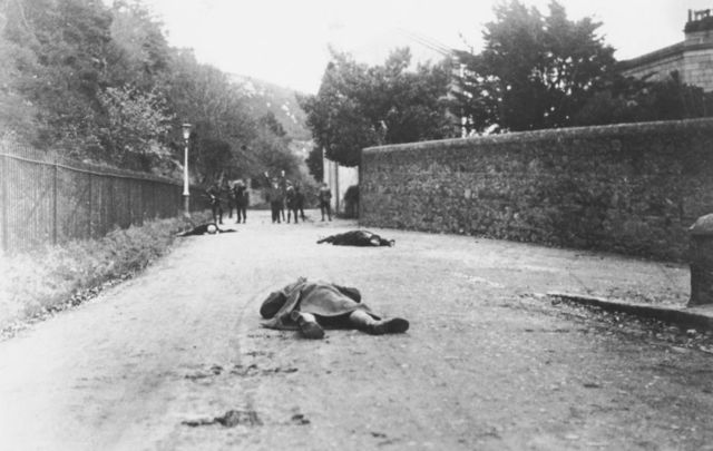 The bodies of dead Sinn Féin supporters lie in the foreground, and in the background, a group of men is arrested by the Royal Irish Constabulary, Ireland, November 1920.