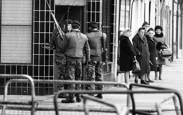Street scene in Belfast during the Troubles. British soldiers stand next to a group of elderly ladies on a street corner. 