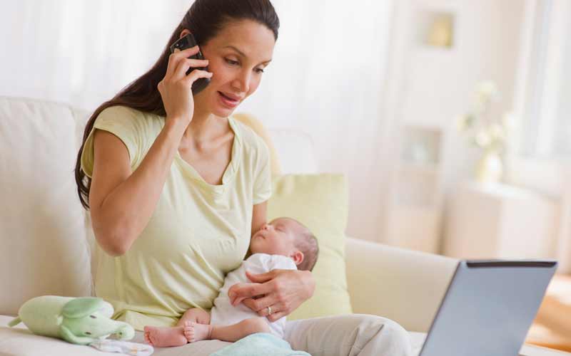 Mother holds newborn baby while talking at phone and looking at computer. Credit: Getty Images